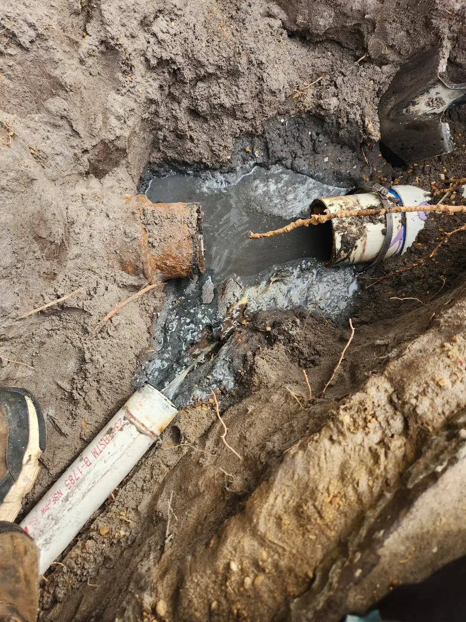 An excavated trench reveals a broken, rusty metal pipe connected to a white PVC pipe, with dirty water pooling around them. Another PVC pipe lies on the ground nearby, and a shovel is visible in the background.