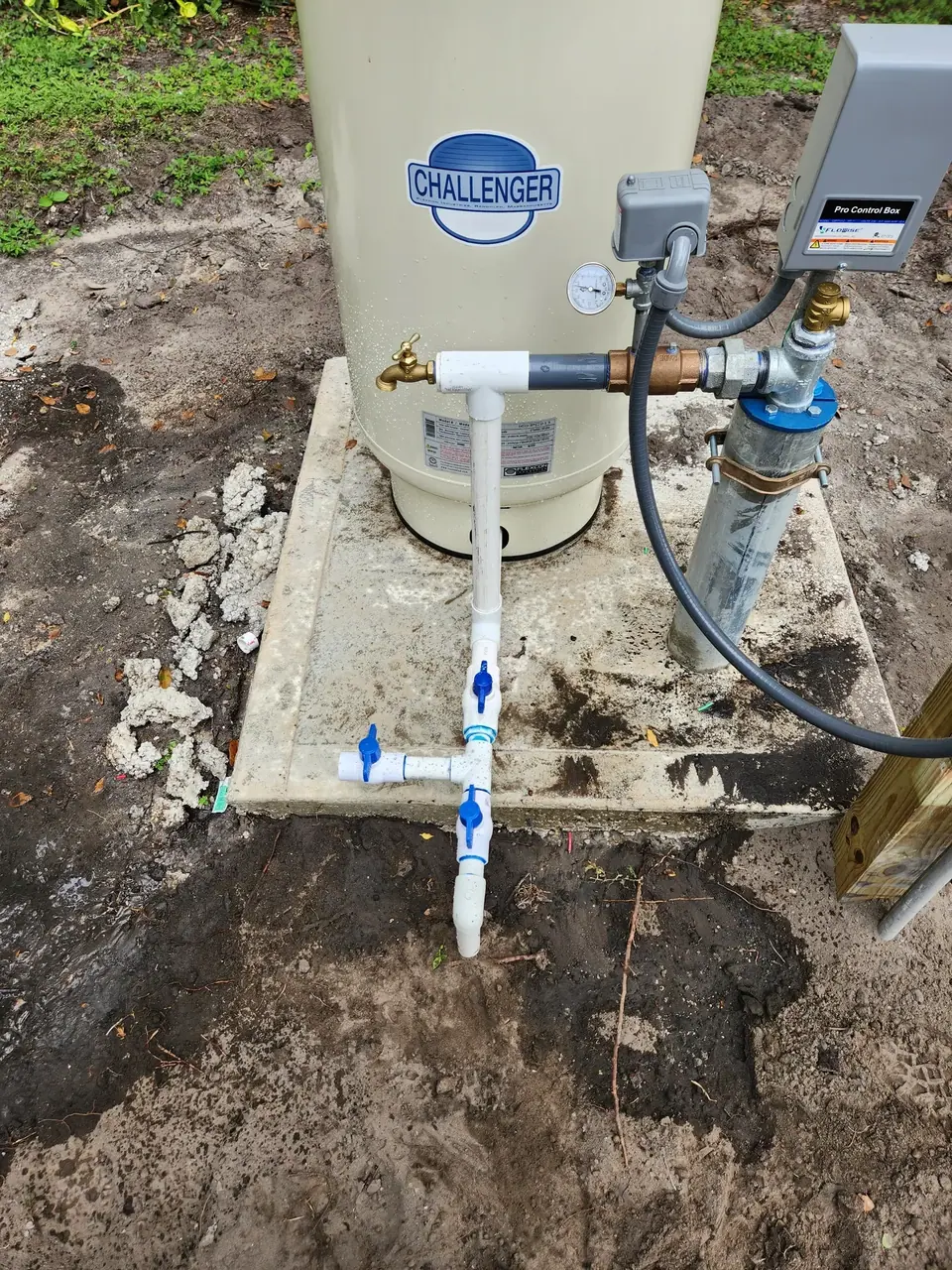 A "CHALLENGER" brand water pressure tank sits on a concrete pad, connected to a well pump system with PVC and metal pipes, blue-handled valves, a pressure gauge, and a "Pro Control Box." The system is installed outdoors on dirt ground with some foliage.