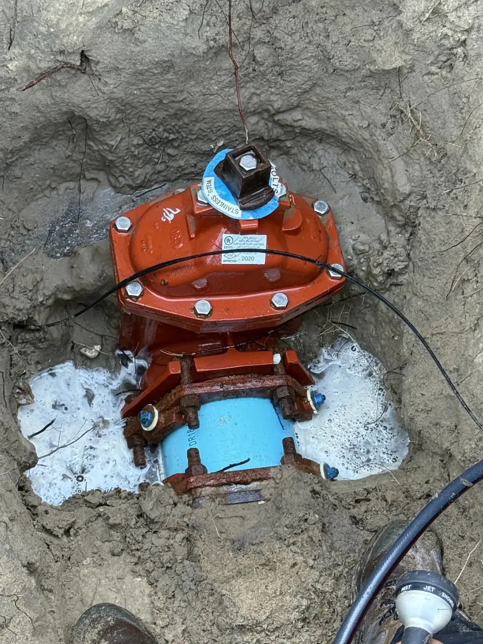 An orange water valve assembly is partially buried in a muddy pit, connected to a light blue pipe at its base. Water with white foam is pooling around the valve, and a black cable runs across its top.