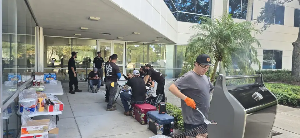 A group of people are having an outdoor cookout under an overhang next to a modern building. A man in a grey shirt and orange gloves is grilling burgers on the right, while others gather around tables with food and supplies.