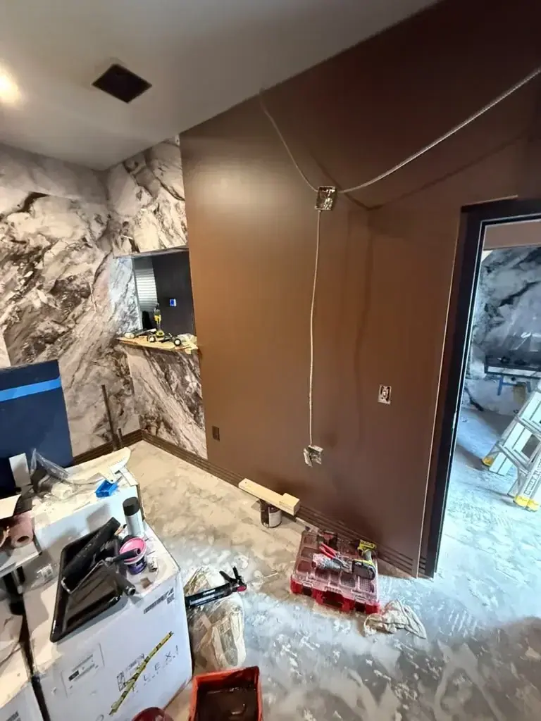 A room under renovation with one wall covered in white and grey marble-patterned material and another painted brown with exposed electrical wiring. Construction tools and materials are scattered on the concrete floor, and a doorway leads to another unfinished area.