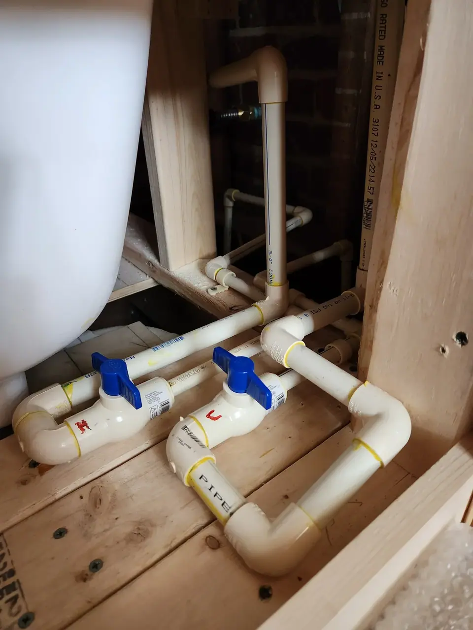 CPVC plumbing pipes with blue shut-off valves are installed on a wooden floor next to a white bathtub, with wooden framing and a brick wall visible in the background.