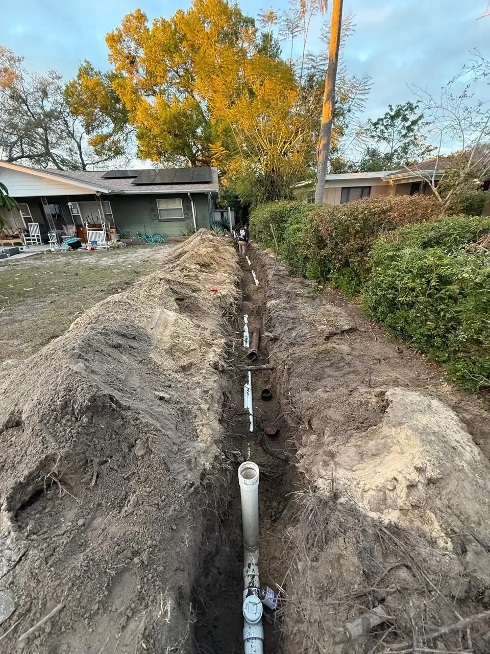 A long trench is dug in a grassy yard, with white PVC pipes laid along its length. Piles of excavated dirt line both sides of the trench, and a house with solar panels is visible in the background.
