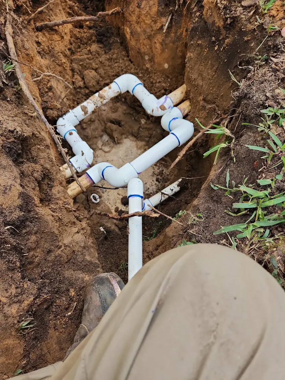 White PVC pipes are laid in a dirt trench, connected with various fittings and showing purple primer. A person's leg and boot are visible in the foreground, looking down at the exposed pipes.