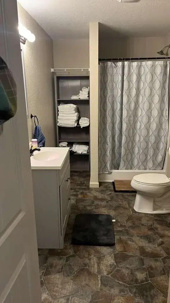 A bathroom features a light gray vanity with a white sink, a dark gray shelving unit stocked with white towels, and a toilet next to a shower with a patterned gray and white curtain. The floor is covered in dark, irregular stone-patterned tiles.