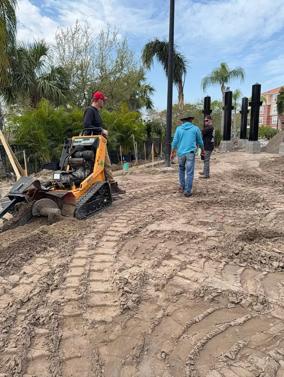 An orange trenching machine is actively digging a trench in a dirt area, with three workers standing nearby. Palm trees and building foundations are visible in the background under a partly cloudy sky.