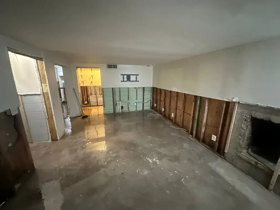 An interior room with a wet concrete floor and stripped-down walls, exposing studs and insulation. The room appears to be undergoing water damage remediation or renovation, with a fireplace opening visible on the right and a TV mount on the far wall.