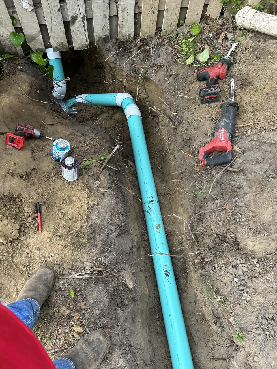 A trench dug in dirt shows teal PVC pipes being installed, with some connected near a wooden fence. Plumbing tools like a reciprocating saw, impact driver, and pipe cement are visible around the trench, along with a person's feet in work boots.
