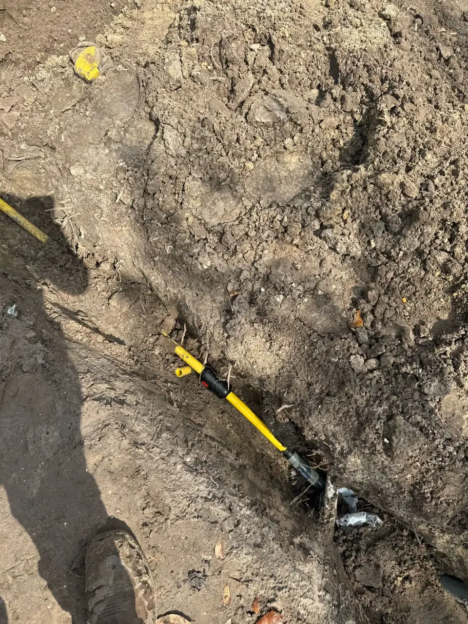 An excavated trench reveals a yellow gas line connected to another pipe underground. A worker's boot is visible in the lower left corner.
