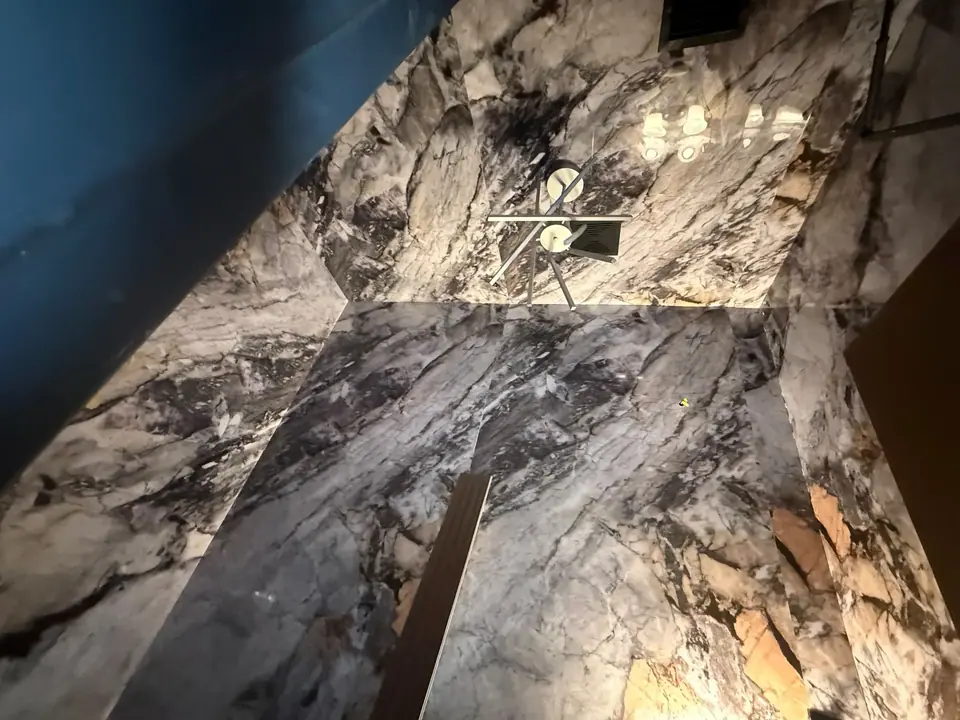 An interior view of a room with walls and ceiling covered in polished, veined marble-like material, featuring grey, white, and dark patterns with some orange tones. A modern chandelier hangs from the ceiling, and a dark blue curved architectural element is visible in the upper left.