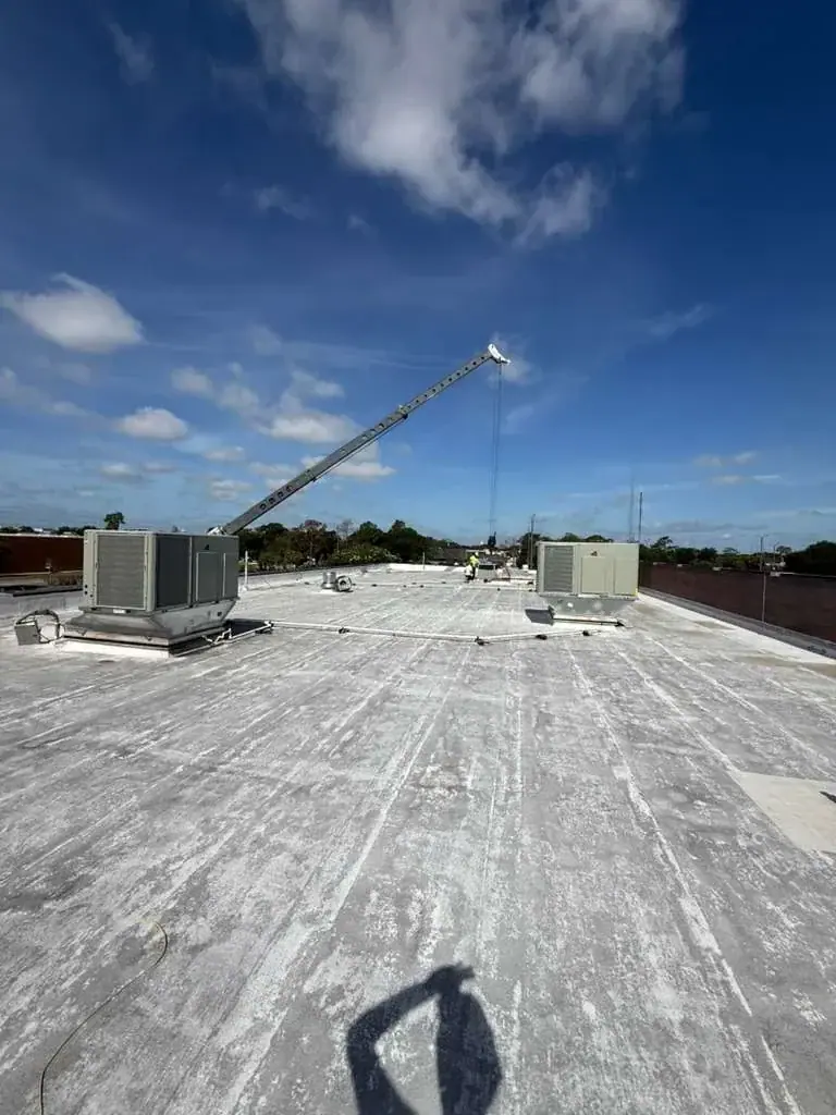 A wide shot of a flat commercial rooftop with two large HVAC units and a crane boom extending into the sky. Workers are visible in the background, suggesting installation or maintenance under a partly cloudy blue sky.