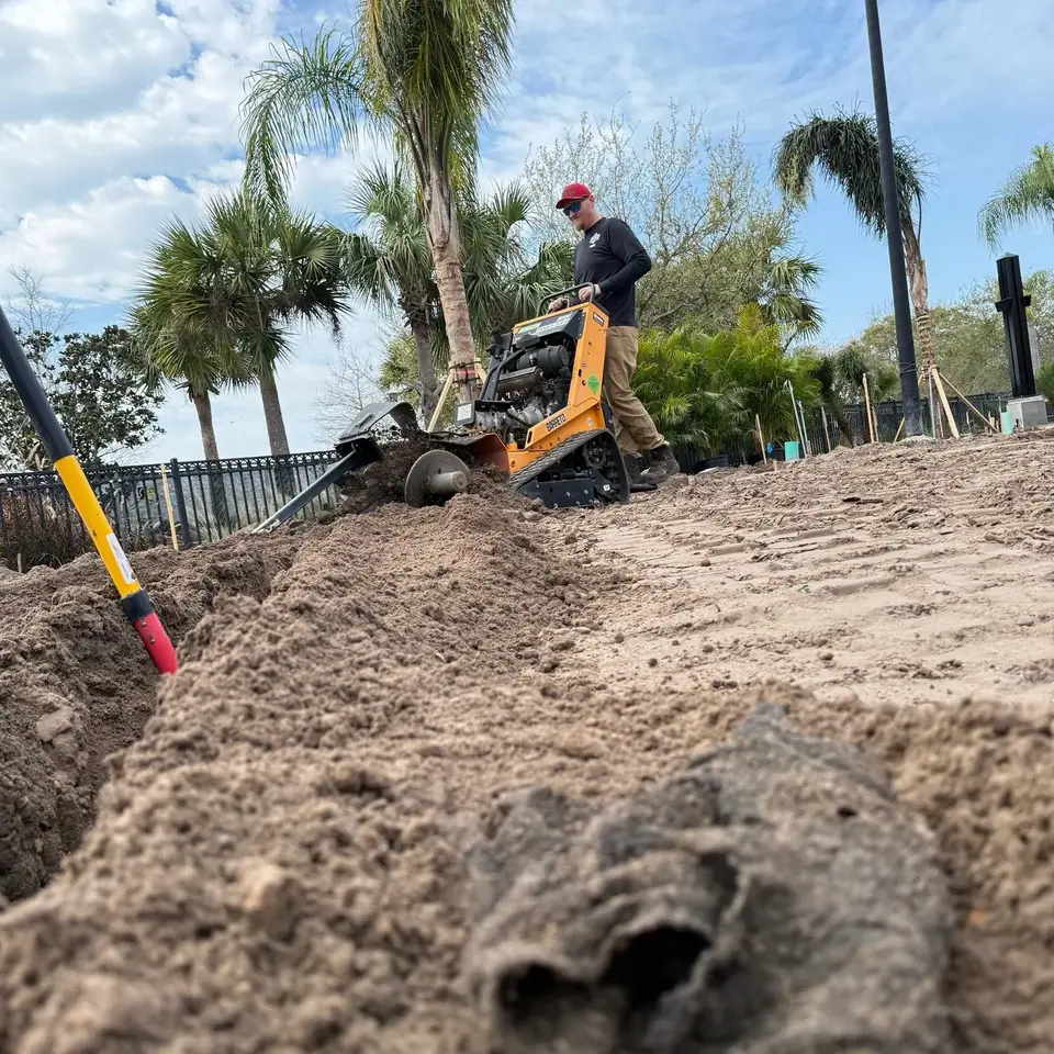 A worker in a red hat and dark shirt operates an orange trenching machine, digging a trench in sandy soil. A yellow and red shovel is stuck in the ground in the foreground, with palm trees and a fence visible in the background under a partly cloudy sky.