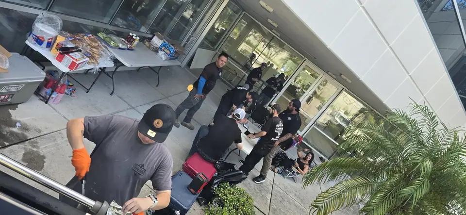 A group of people, mostly men, are gathered outdoors near a building with large glass windows. Two tables are set up with food items like buns and condiments, and one man in the foreground wears orange gloves while working on a metal object.