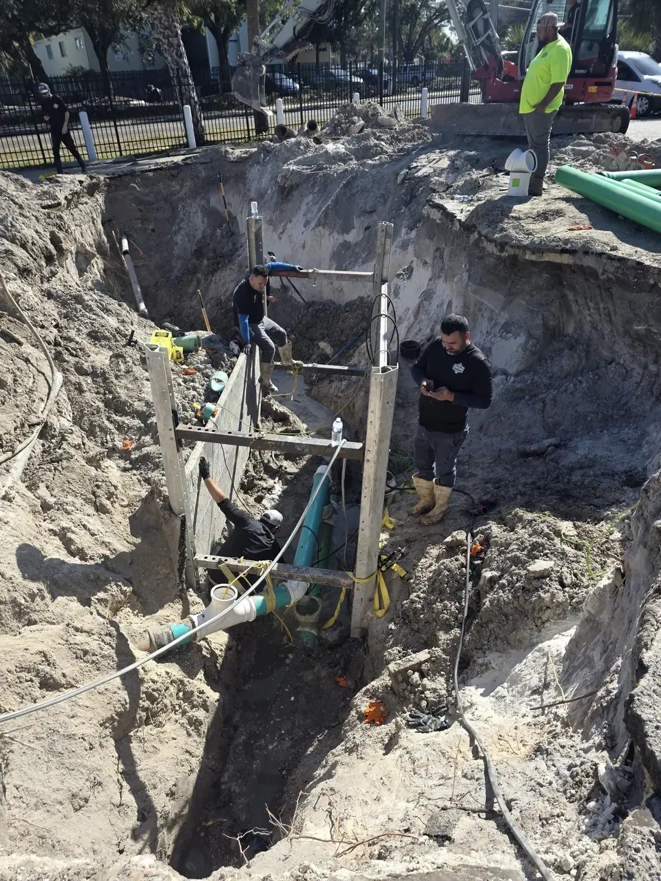 A construction site features a deep, wide trench where several workers are installing or repairing pipes. An excavator is visible at the top edge of the trench, with workers at various depths within the excavation.