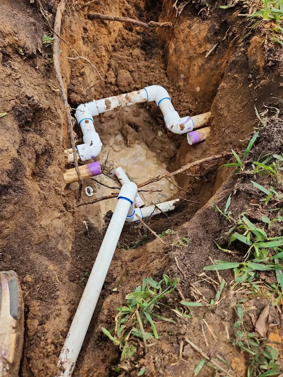 A trench dug in brown dirt reveals white PVC pipes, some connected with elbows and couplings, lying in muddy water. Tree roots are visible along the sides of the trench.