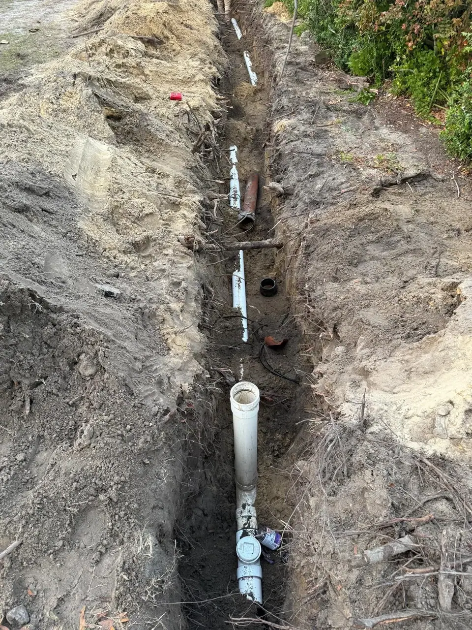 A long, narrow trench is dug into the dirt, revealing several white PVC pipes and one reddish-brown pipe laid along its length. Piles of excavated soil are visible on both sides of the trench, with some roots and debris scattered around.