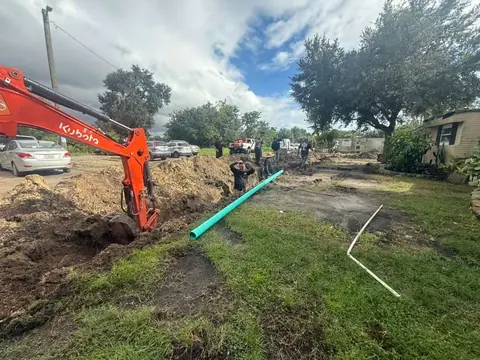 An orange Kubota excavator digs a trench in a grassy residential area, with several workers, a large dirt pile, and a long light blue pipe laid out beside it.