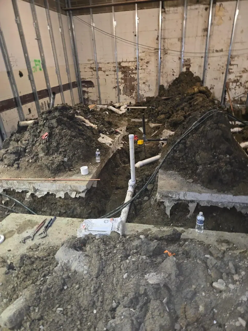 An indoor construction site shows trenches dug into the floor with new white PVC pipes and copper lines being installed. Piles of excavated dirt are on either side of the trenches, and the background features unfinished walls with metal studs.