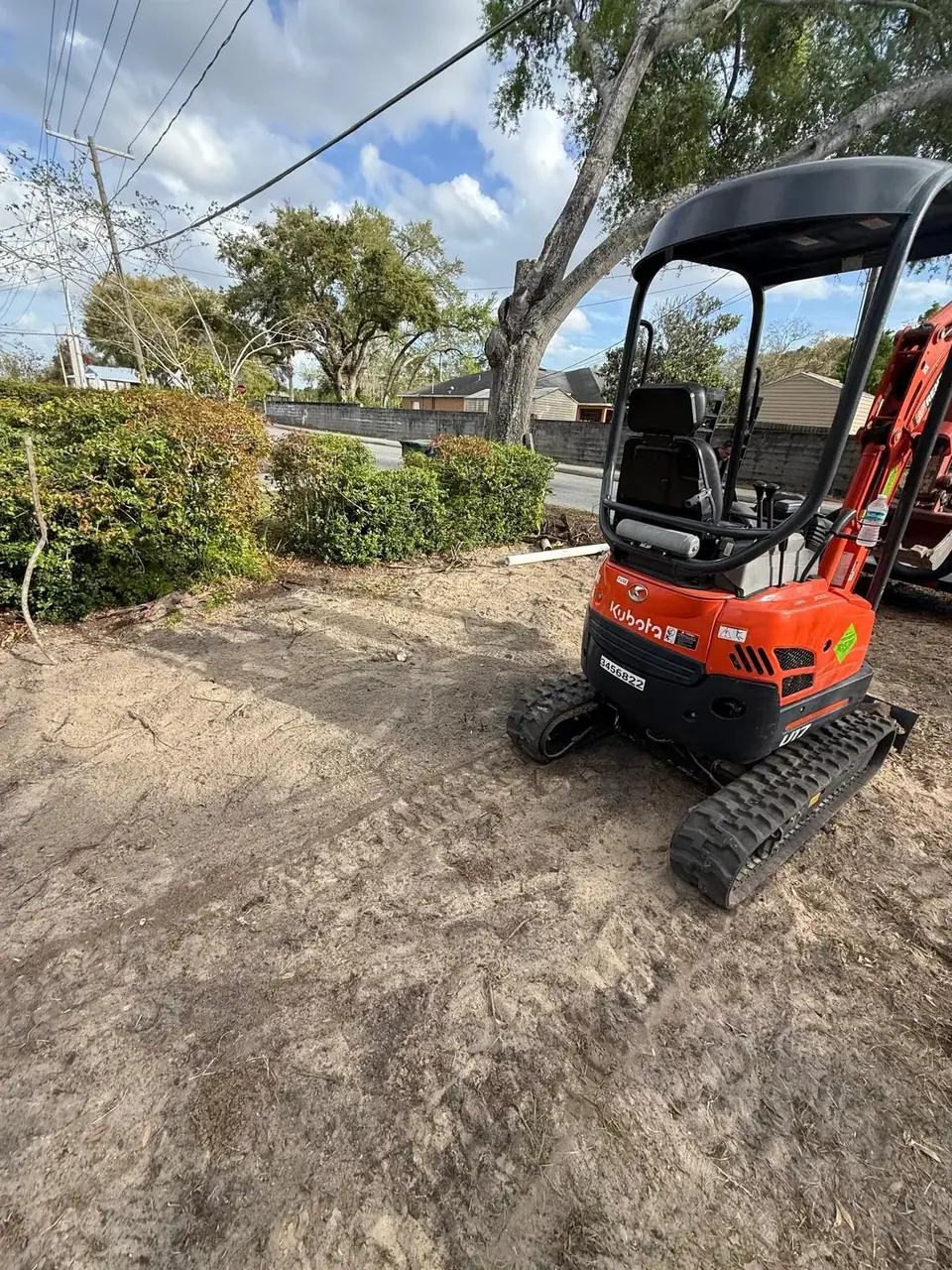 A bright orange Kubota mini excavator is parked on a patch of sandy dirt next to a green hedge. In the background, a street, power lines, and residential houses are visible under a partly cloudy sky.