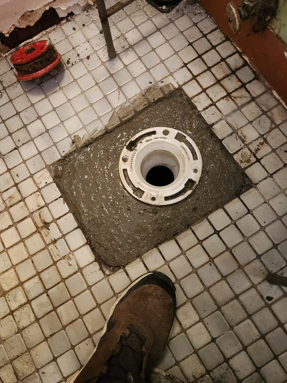 A white toilet flange is set into wet concrete on a dirty, light-tiled floor, with a brown shoe visible in the foreground. A red plumbing test plug and damaged wall are in the background.
