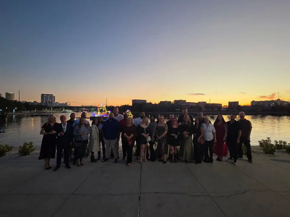 A group of approximately 25 people stands on a paved waterfront promenade at dusk, with a city skyline and a body of water reflecting sunset colors and lights in the background.