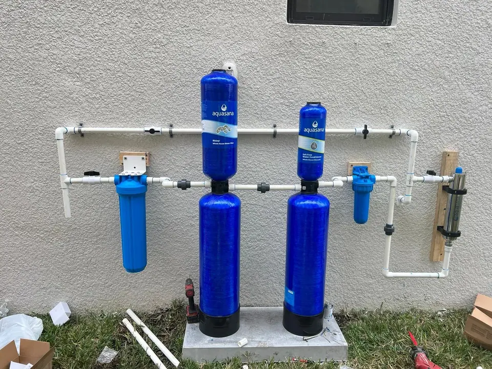 A whole-house water filtration and conditioning system is installed against a textured white wall, featuring two large blue Aquasana tanks, two smaller blue filter housings, and a silver UV filter. White PVC pipes connect the components, which are mounted on a concrete pad with grass and construction debris visible below.
