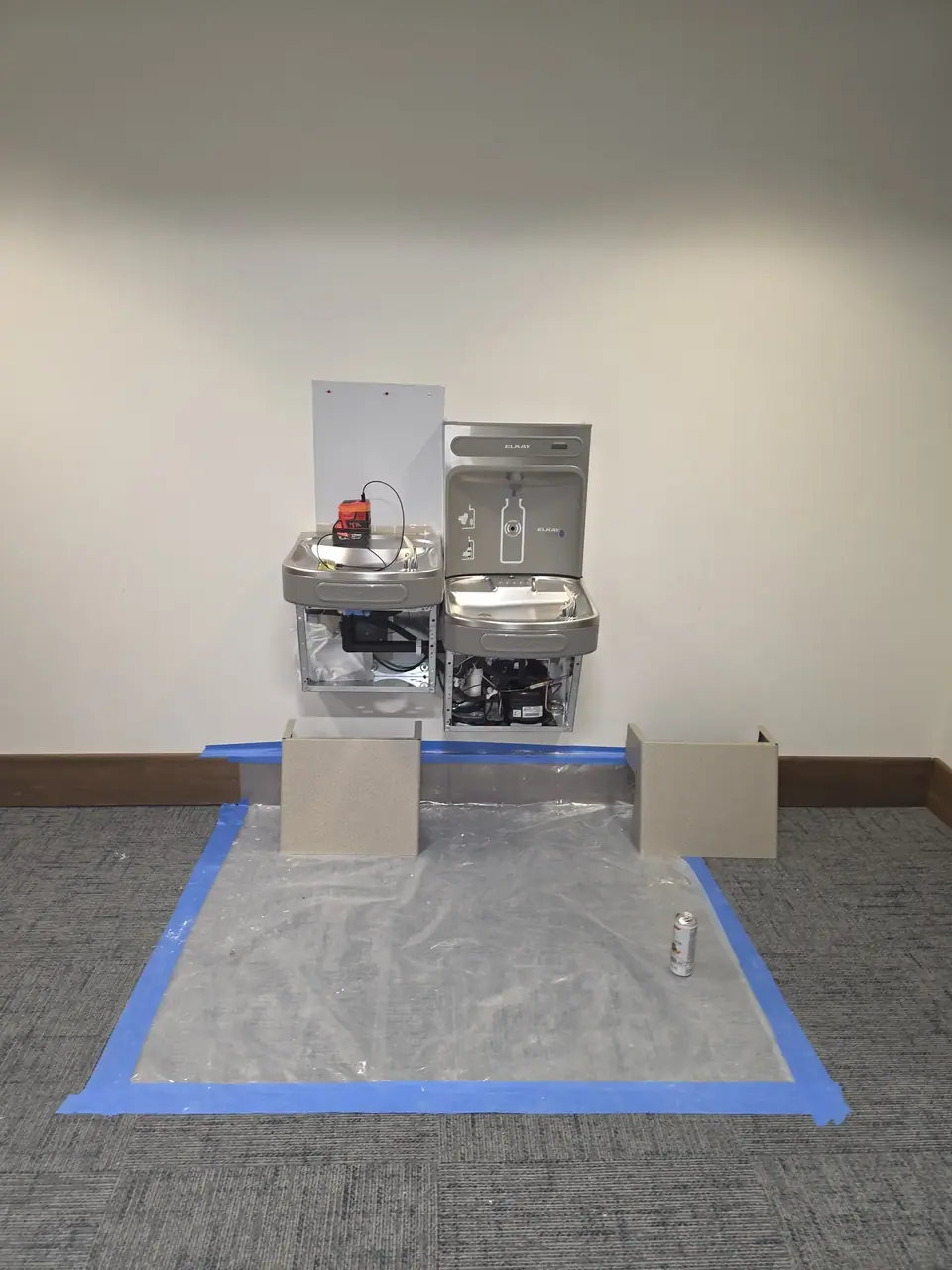 Two Elkay water bottle filling stations are mounted on a white wall, with their lower panels open revealing internal components. A red and black device sits on the left unit, and a plastic sheet is taped to the carpeted floor below, with two beige panels and a soda can on it.