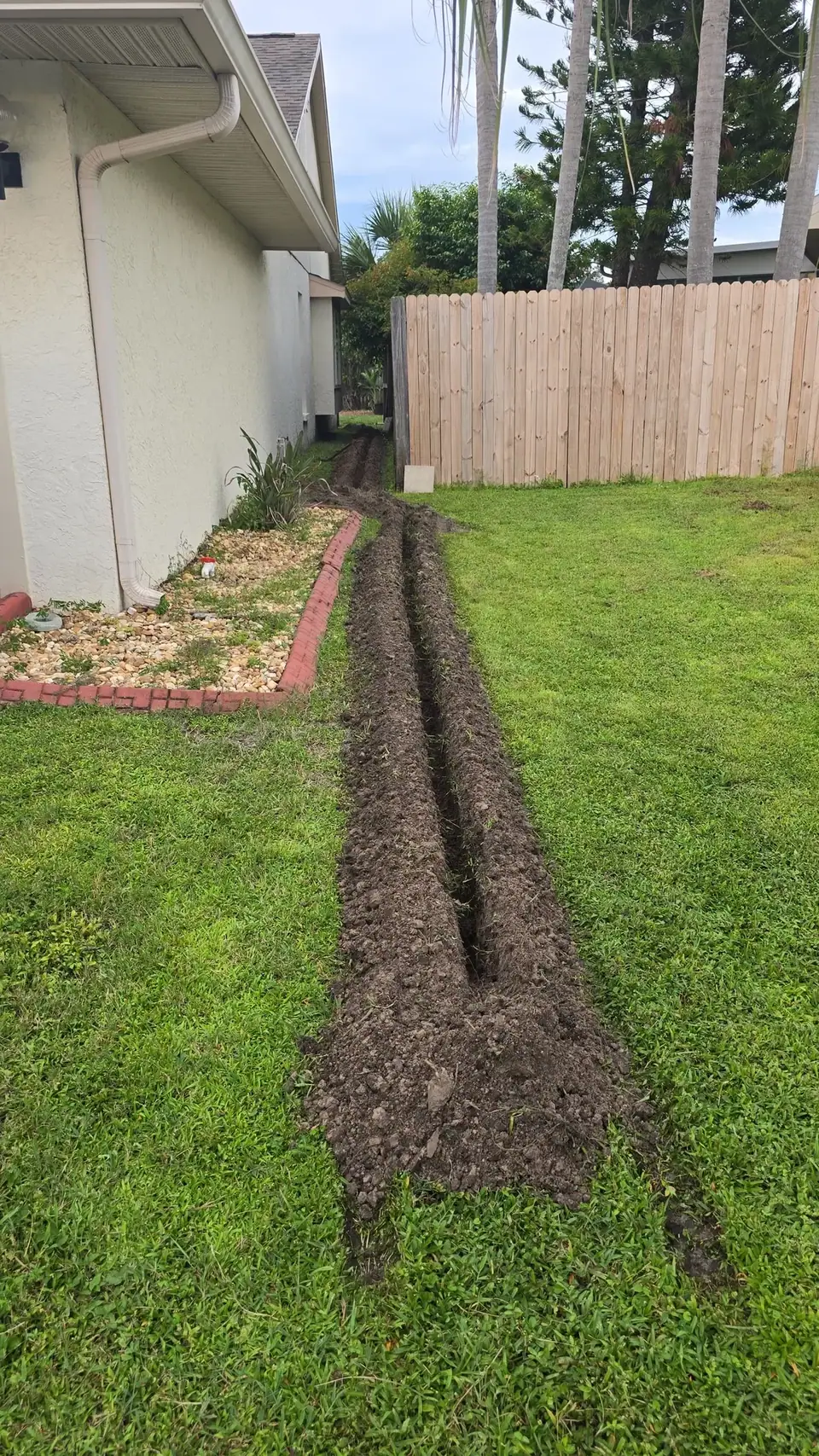 A long, narrow trench with two parallel channels and excavated soil runs through a green grassy backyard. It extends from alongside a house with a downspout towards a wooden fence and palm trees in the background.