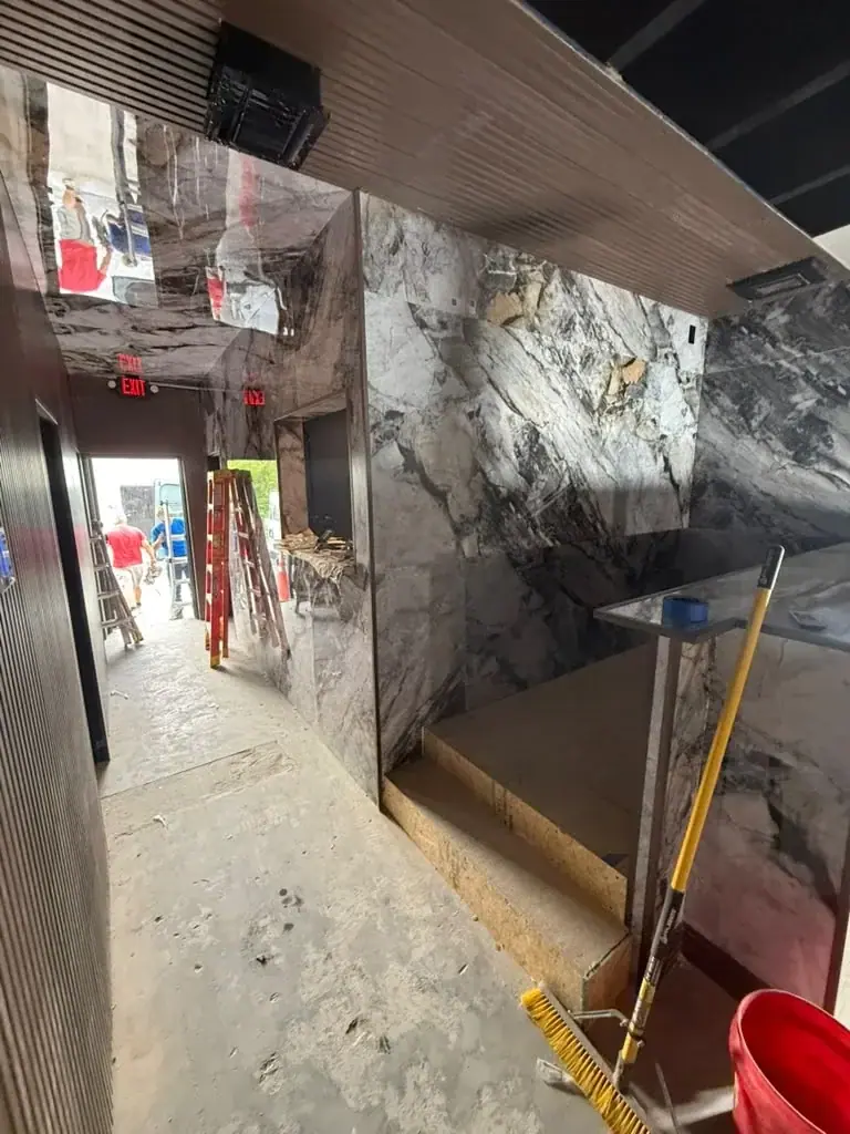 An interior space under construction features marble-patterned walls, an unfinished concrete floor, and wooden steps leading to a counter. Construction tools like ladders and a broom are present, with people visible through a doorway in the background.