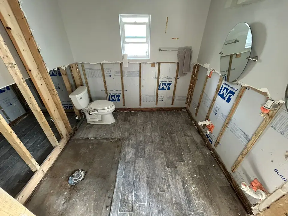An interior view of a bathroom under renovation, showing exposed wall studs, insulation boards, a toilet, a window, a mirror, and a floor with a mix of concrete and wood-look tiles.