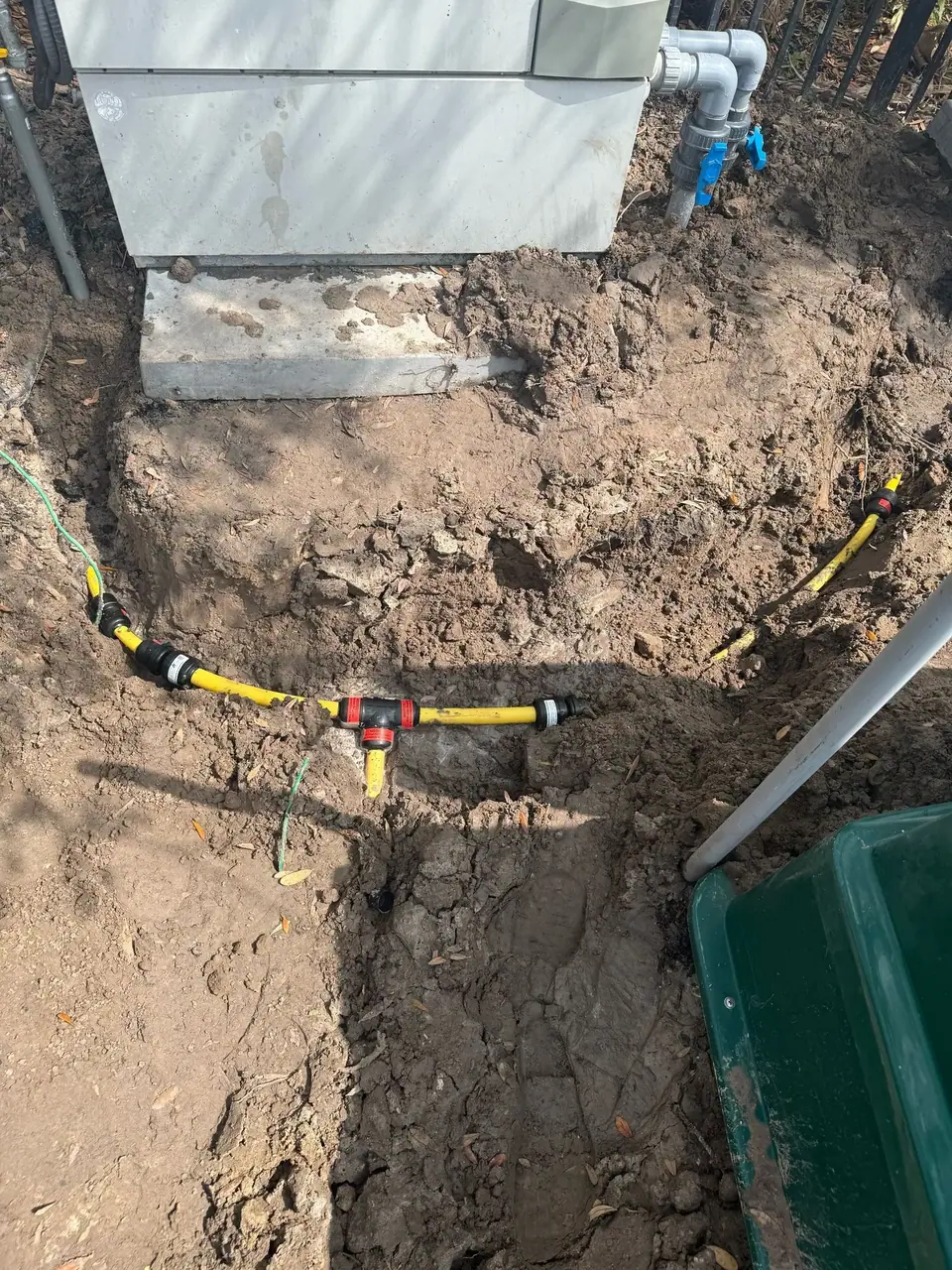 The photo shows yellow flexible pipes with black connectors laid in a dirt trench next to a large grey unit on a concrete slab, with grey pipes connecting to the unit. A green wire is also visible in the trench.