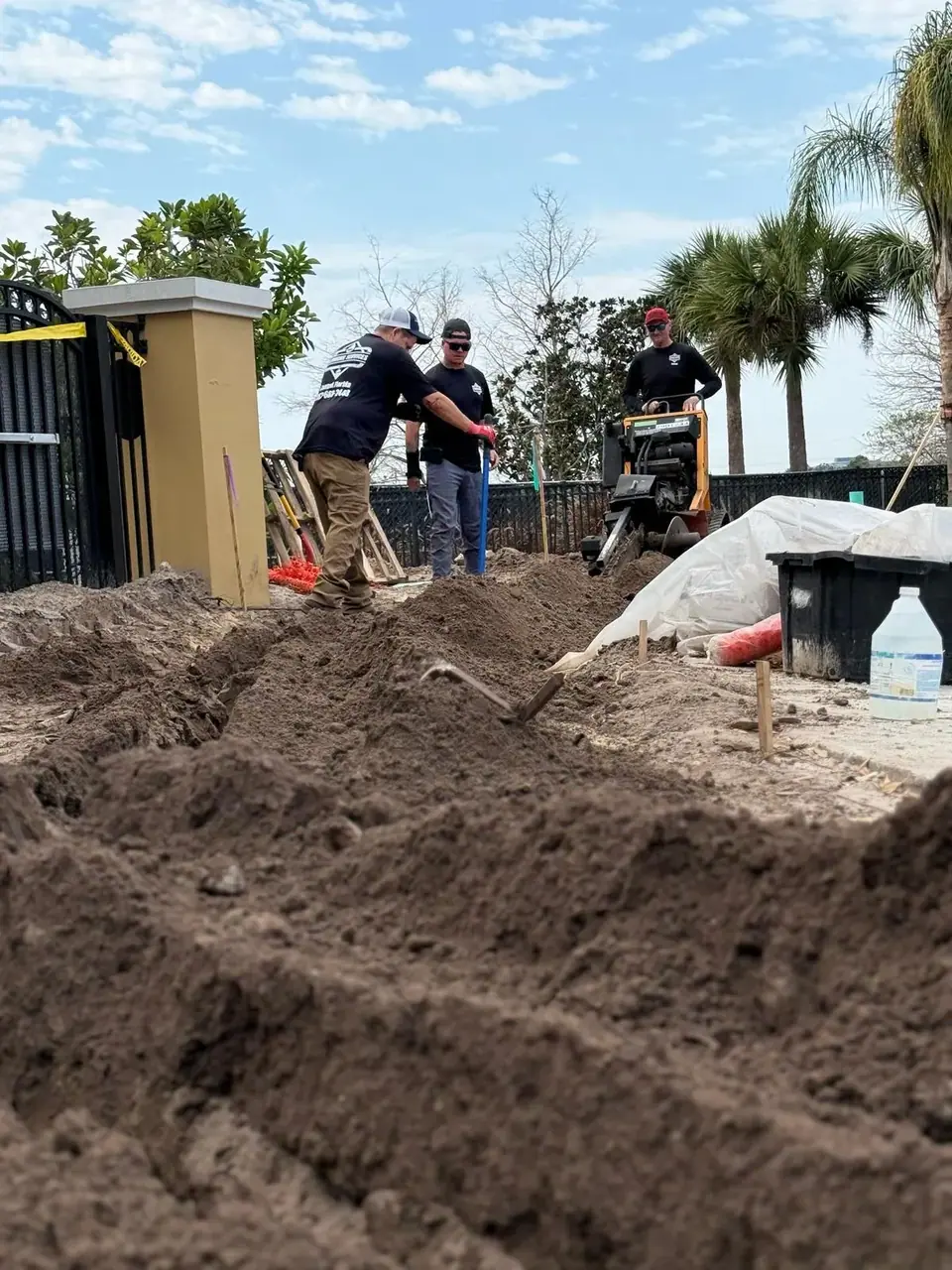Three plumbers are working outdoors on a trenching project. One worker operates a trenching machine while two others stand nearby, with piles of dirt flanking a newly dug trench.