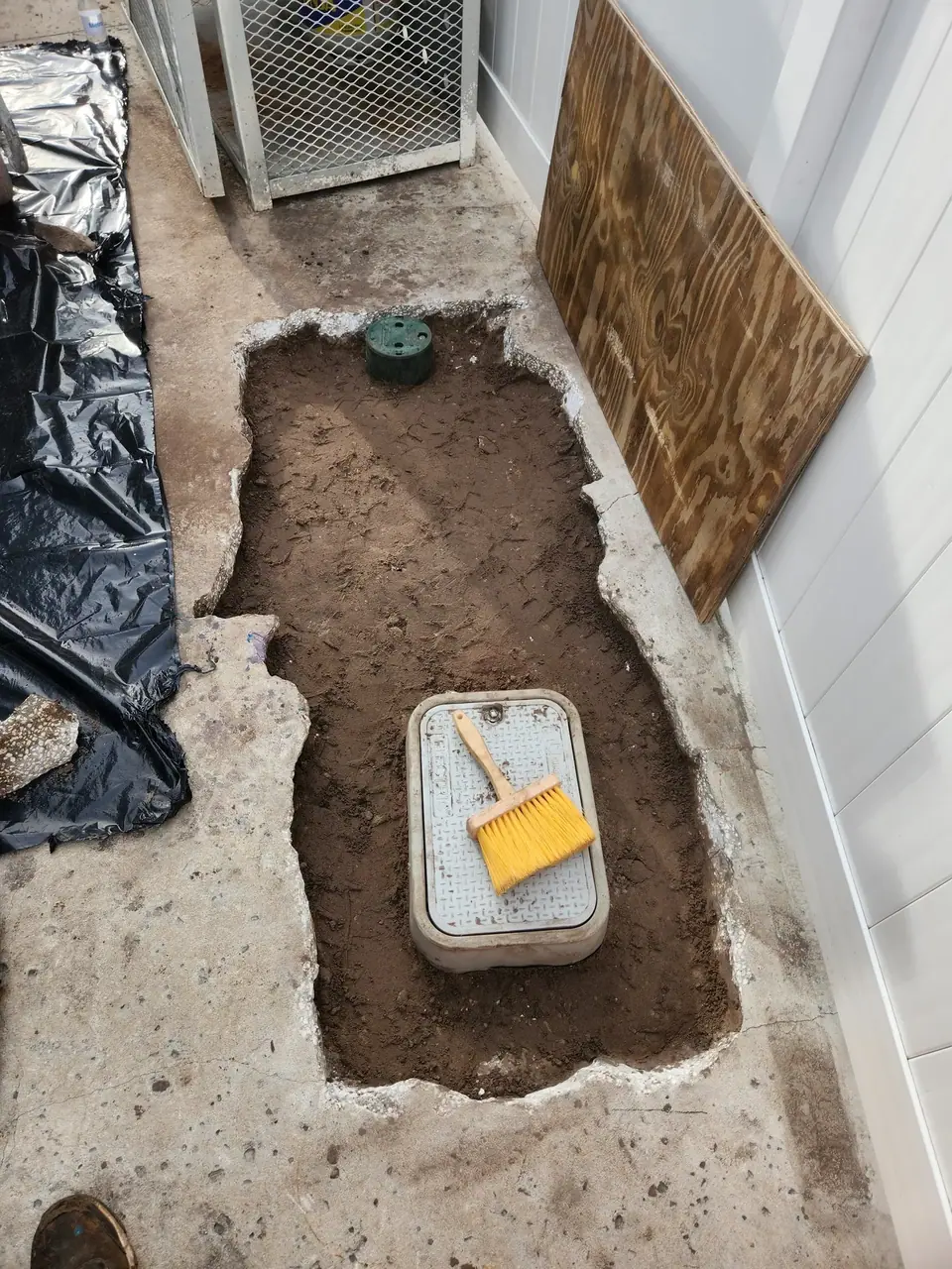 An excavated area in concrete reveals dirt, a grey utility box with a yellow brush on top, and a smaller green cap. Black plastic sheeting is on the left, and a piece of plywood leans against a white wall on the right.