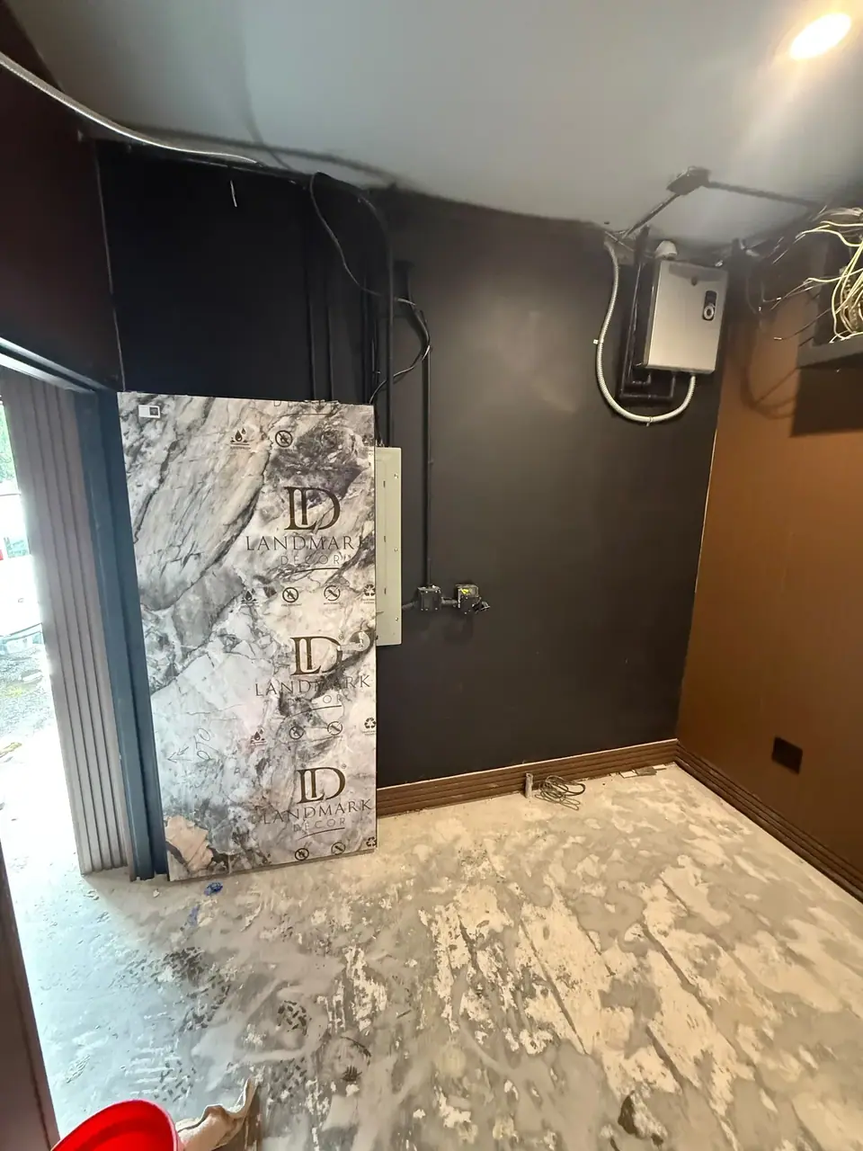 An indoor space under renovation with a large marble-patterned sheet labeled 'LANDMARK DECOR' leaning against a dark wall. The concrete floor shows remnants of removed flooring, and exposed wiring and an electrical box are visible on the walls.