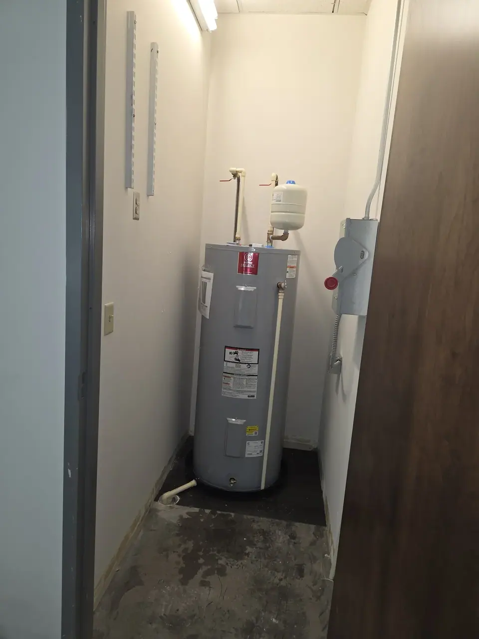 A gray water heater with an expansion tank is situated in a small utility closet with white walls and a concrete floor. An electrical disconnect box is mounted on the right wall, and shelving tracks are visible on the left wall.