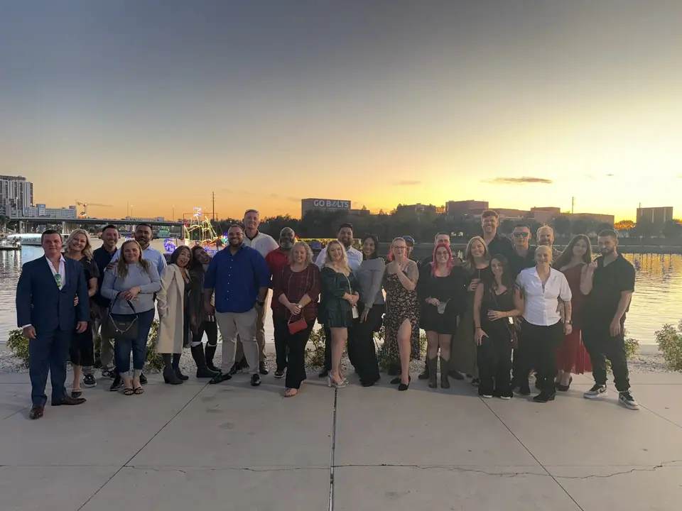 A large group of diverse adults stands outdoors on a paved area at sunset. In the background, a body of water reflects the orange sky, with city buildings, a 'GO BOLTS' sign, and a decorated boat visible.
