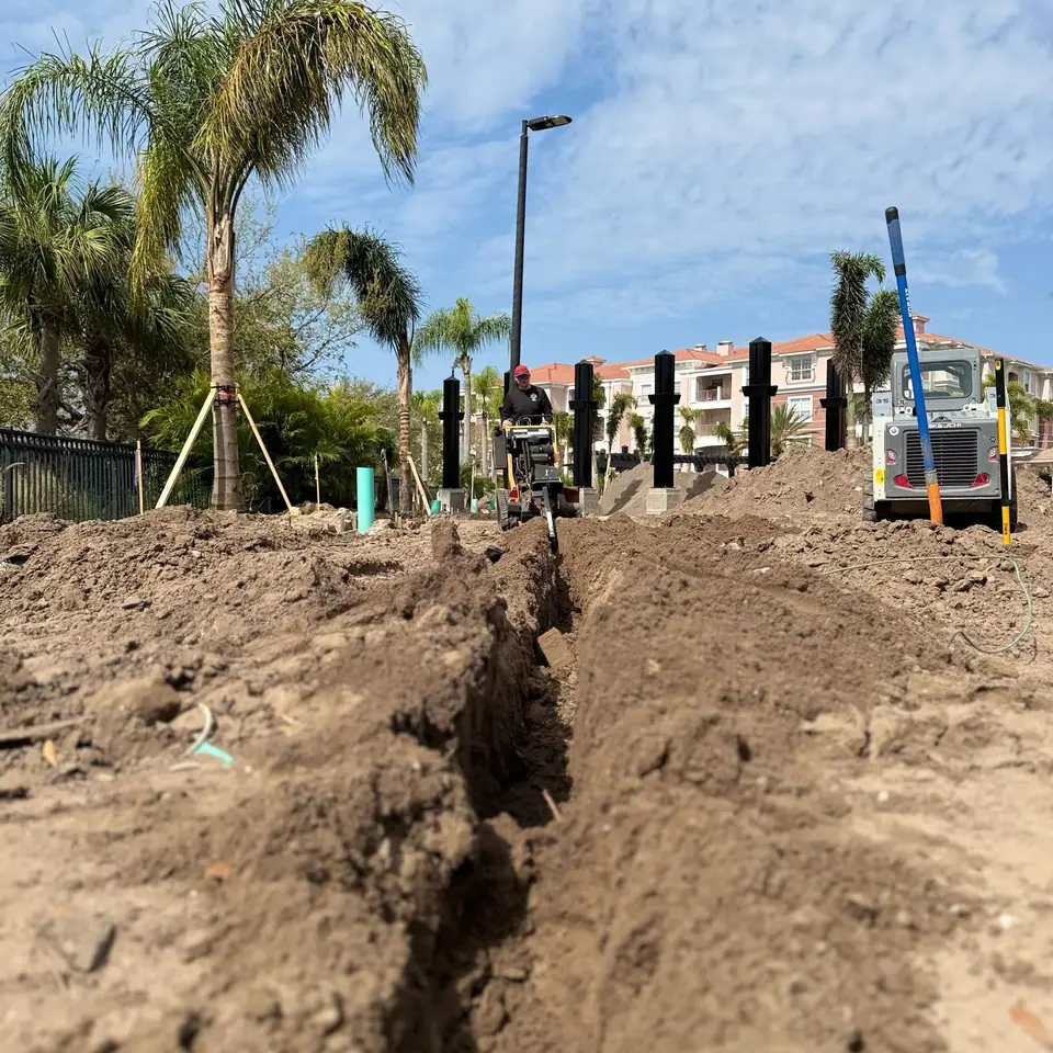 A worker operates a trenching machine, digging a narrow trench through a dirt-covered area. Piles of excavated soil line the trench, with palm trees and buildings visible in the background under a partly cloudy sky.