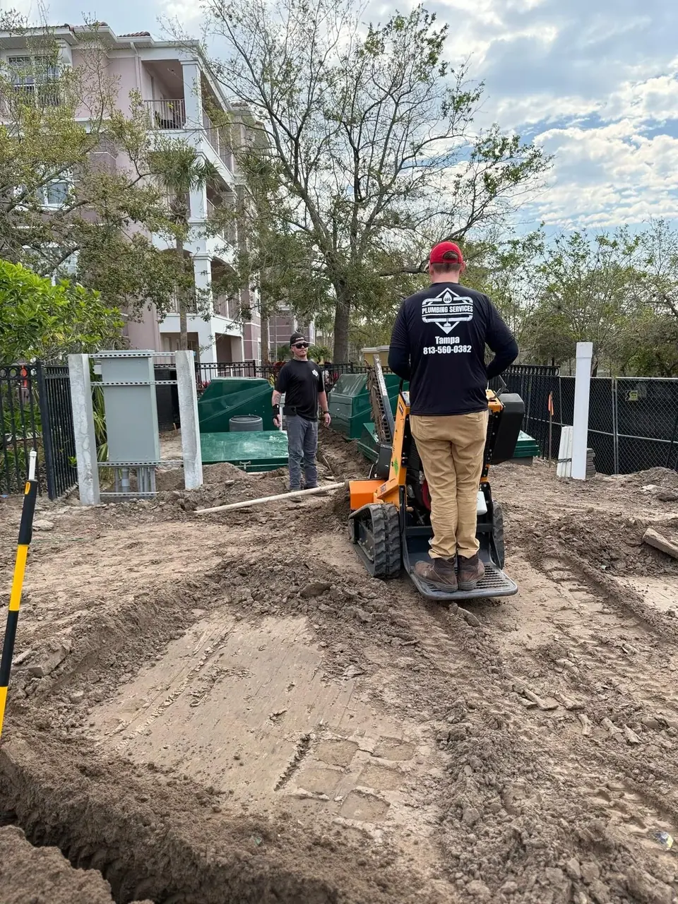 Two men are at a construction site with dug-up dirt. One man operates a small tracked machine, while the other stands near green tanks, with a multi-story building in the background.