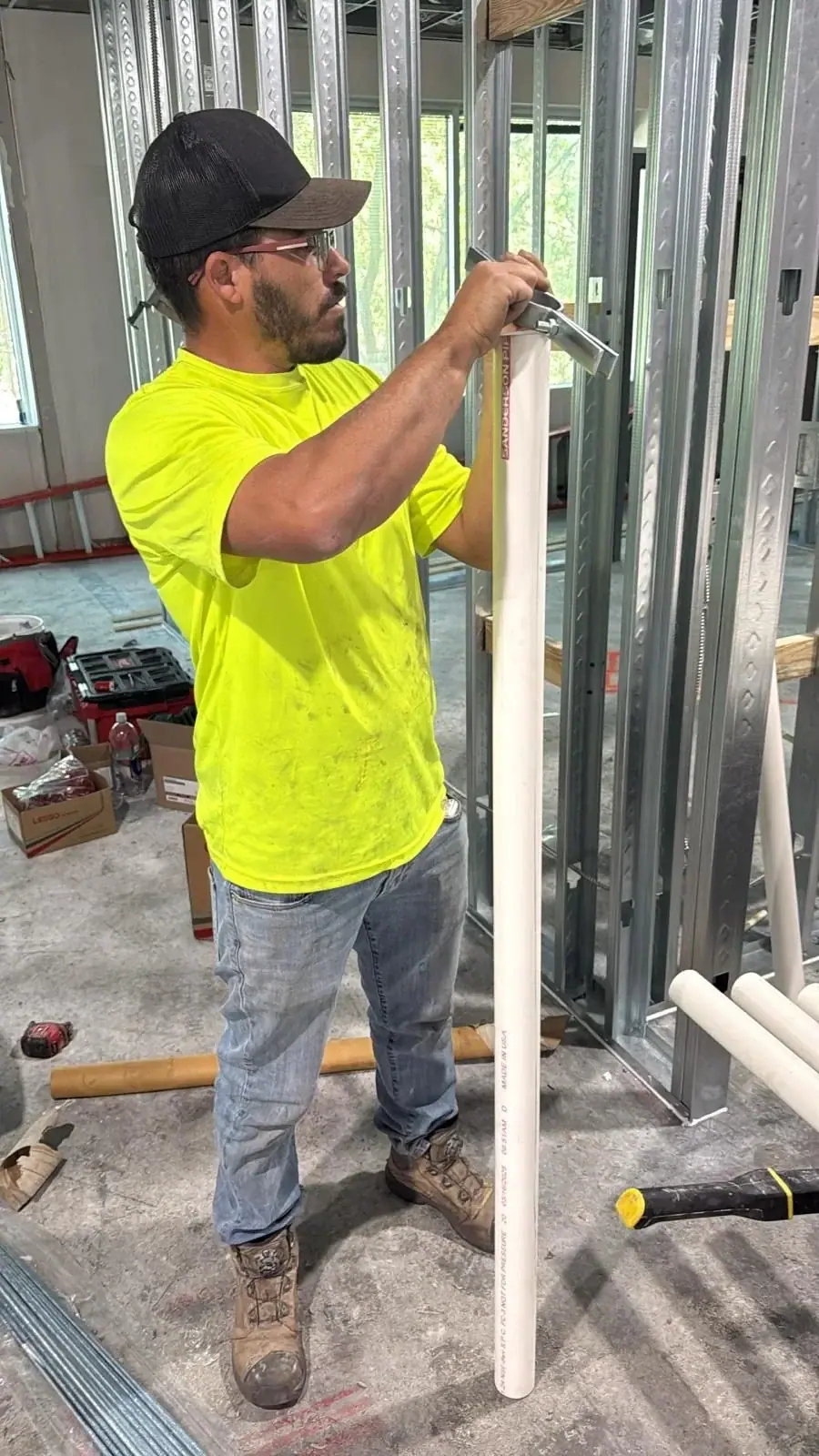 A worker in a neon yellow shirt, jeans, and a black cap is shown from the waist up, holding a white PVC pipe and a tool, working amidst metal wall studs on a construction site.