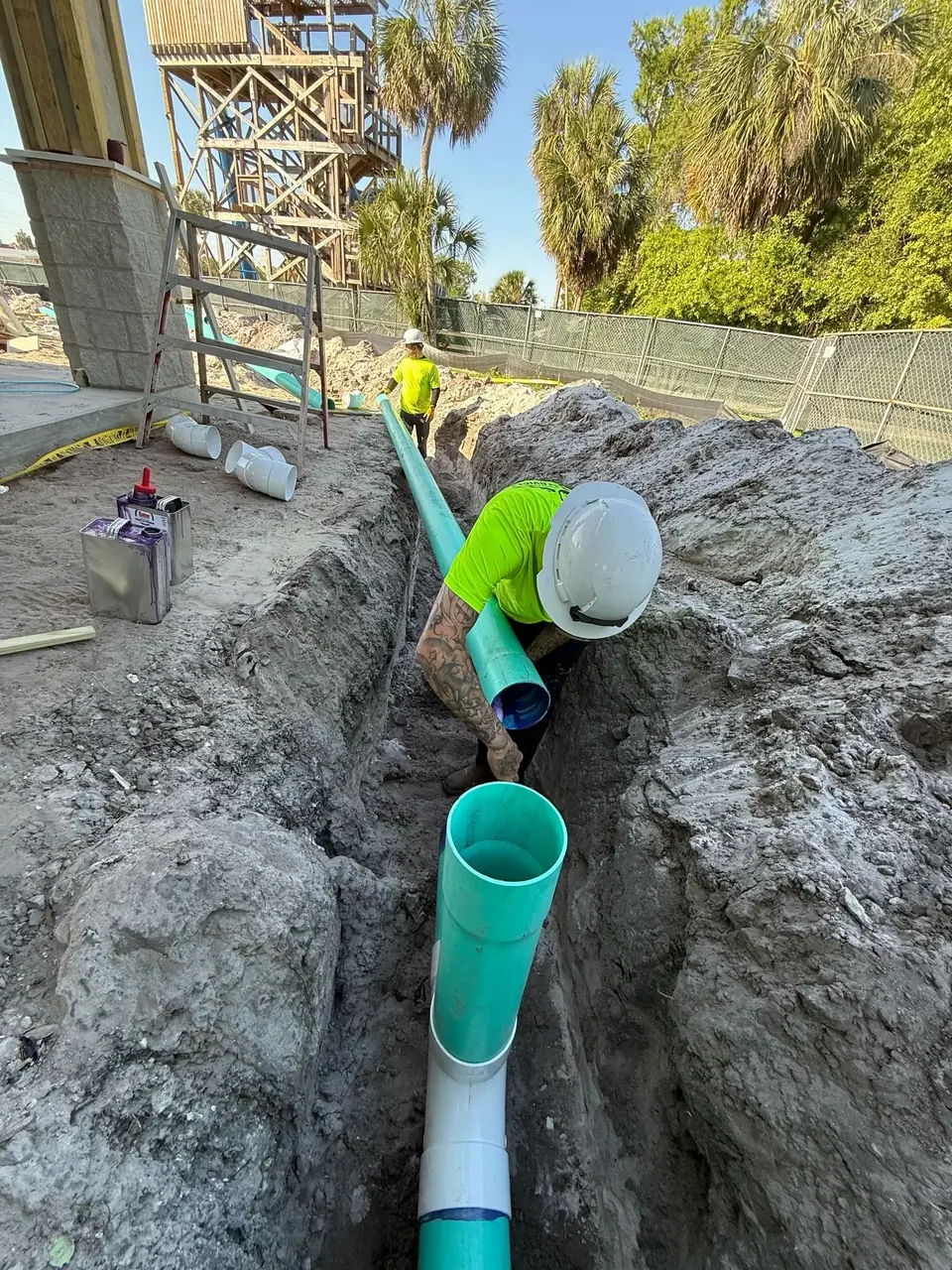 A construction worker in a neon shirt and hard hat is in a dirt trench, connecting a green pipe to another pipe. Another worker stands in the background near a large wooden structure, with palm trees and a chain-link fence visible under a clear sky.