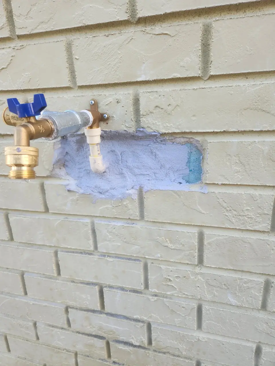 A brass outdoor water spigot with a blue handle is installed on a light yellow painted brick wall. A section of the brick around the spigot is cut out, revealing a light blue material and grey sealant.