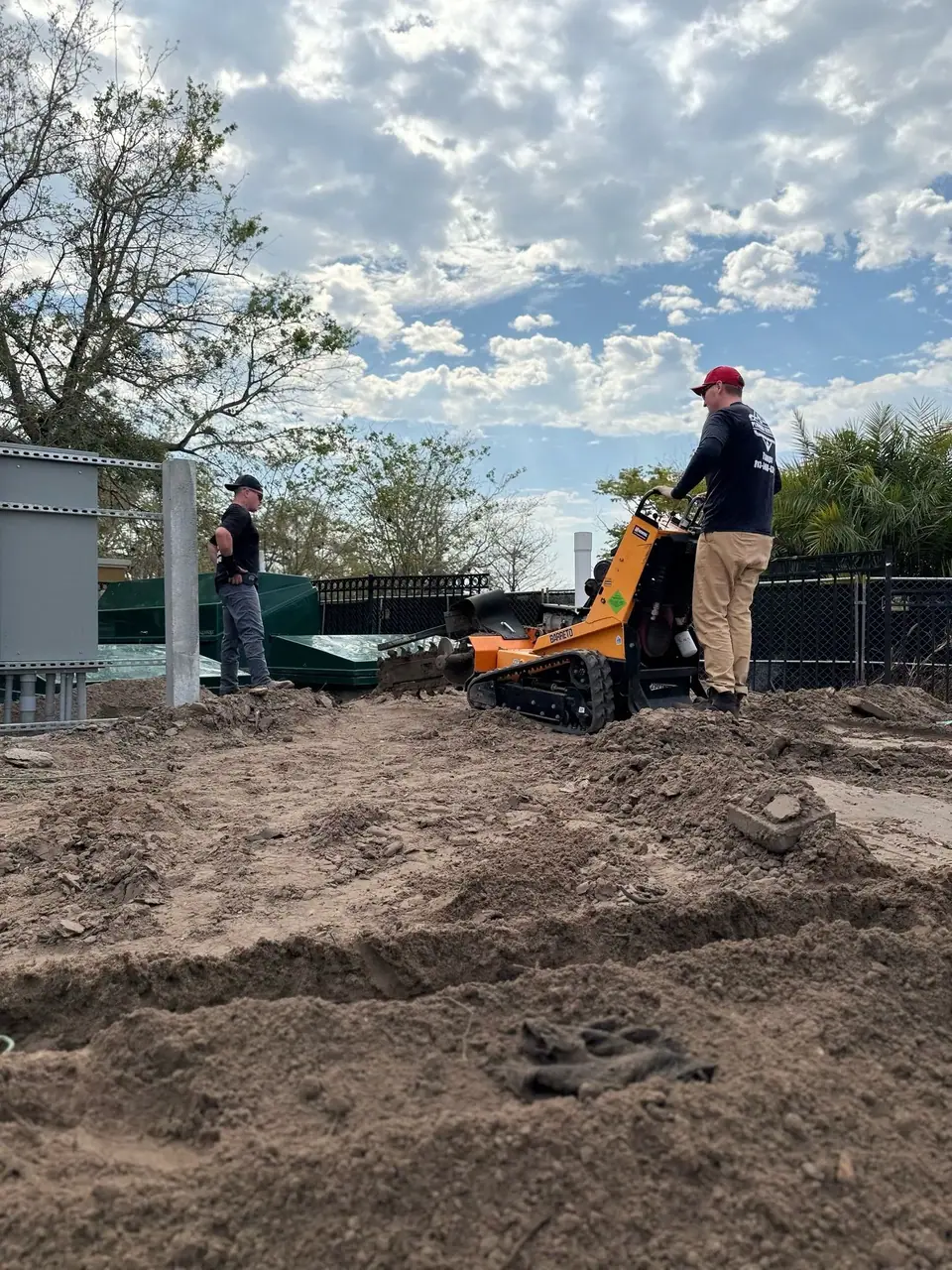 Two men are working on a construction site with loose dirt. One man operates an orange tracked machine, while the other stands near a concrete pillar and green structures under a cloudy sky.