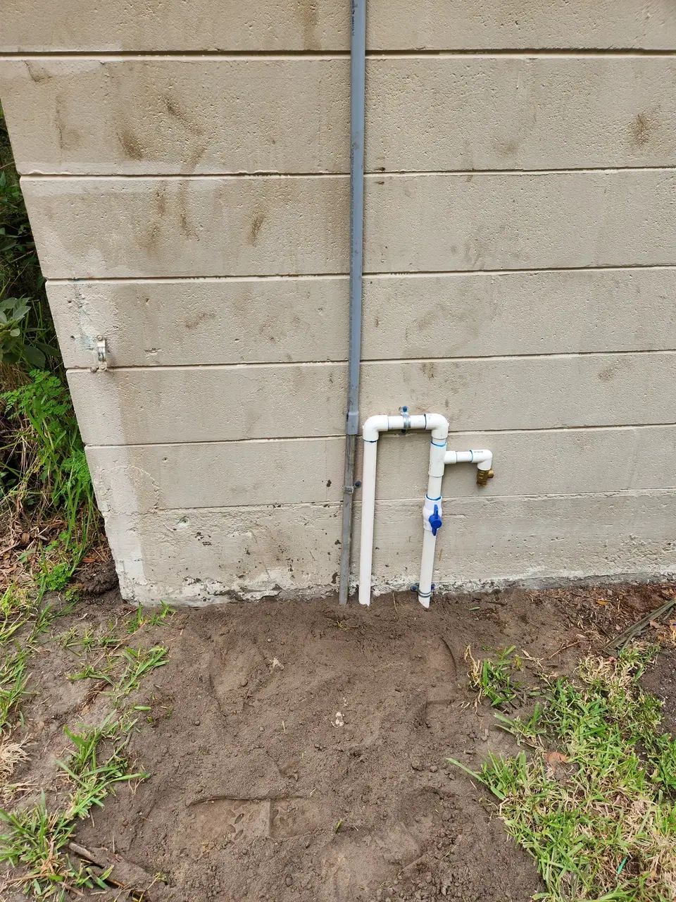 A light-colored concrete block wall with a grey vertical pipe and a white PVC pipe assembly attached, featuring a blue valve and a brass spigot. The ground below is dirt with patches of green grass.