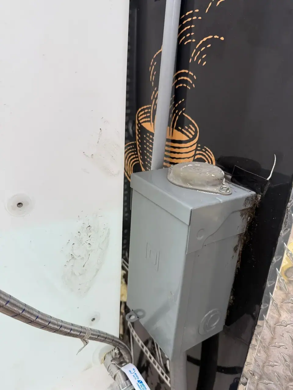 A close-up view of a grey electrical box with a conduit, mounted between a white wall and a black surface with orange patterns. A braided hose and valve are visible at the bottom left.