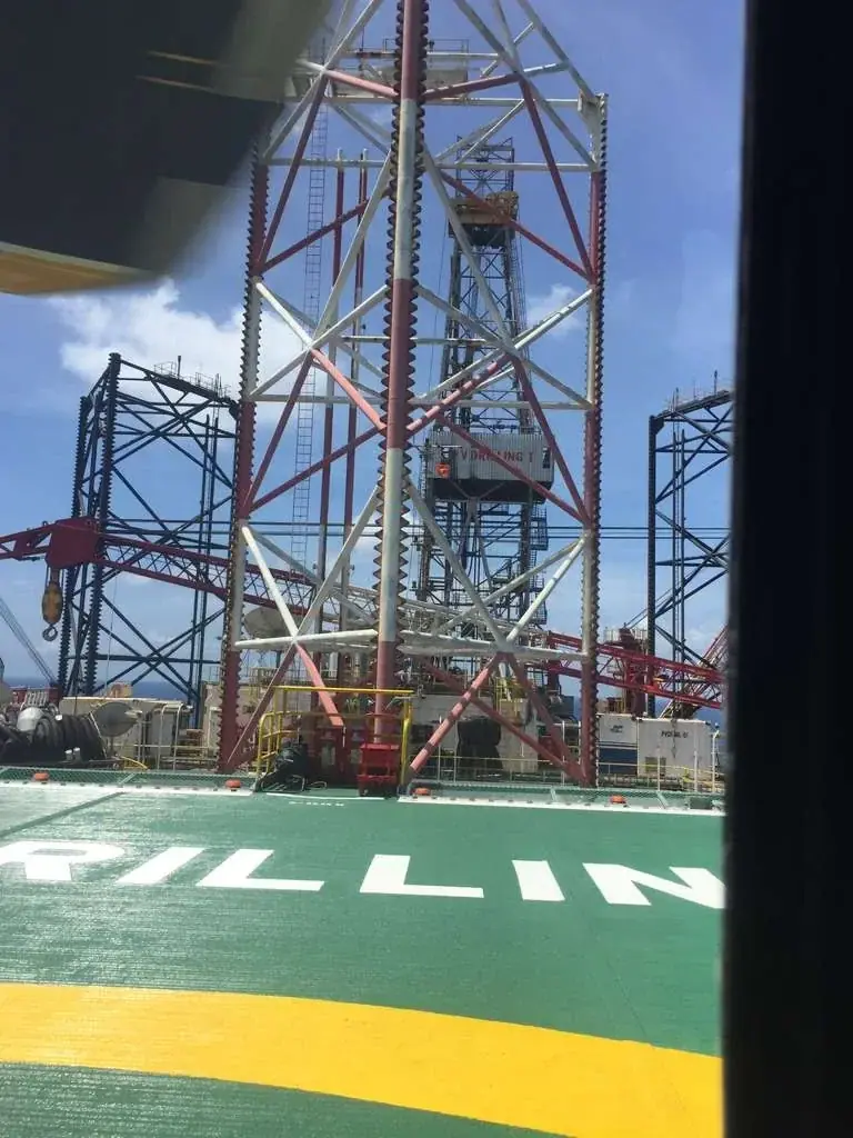 A view from a helipad on an offshore drilling rig, showing the tall red and white derrick structure against a blue sky. The green helipad in the foreground has white letters partially visible, spelling 'DRILLING'.