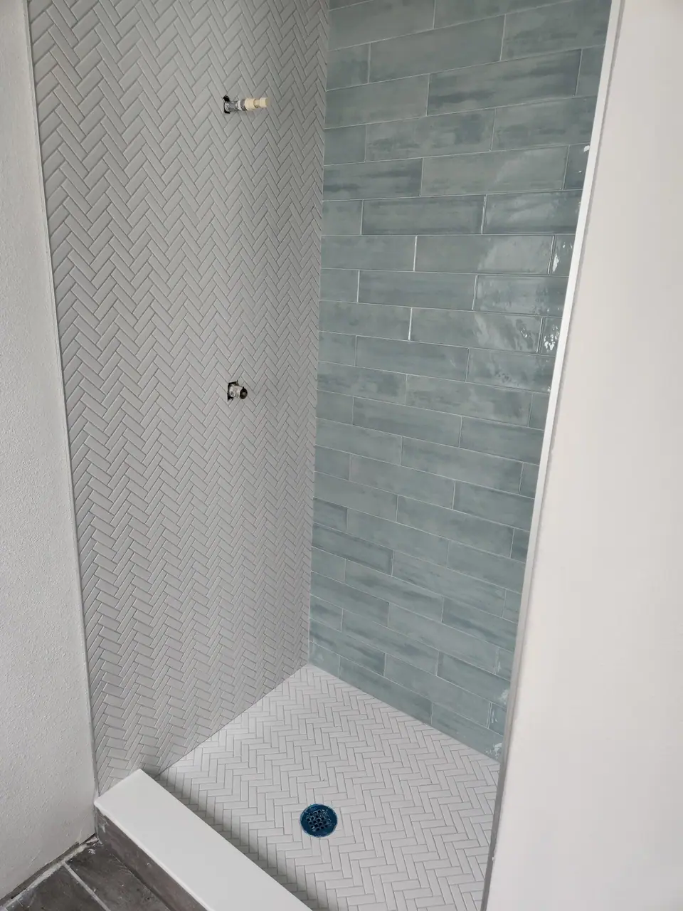 A shower stall under construction features light gray herringbone tiles on the left wall and floor, and blue-green rectangular tiles on the back wall. Exposed plumbing for a shower head and faucet are visible on the left wall, with a blue drain on the floor.