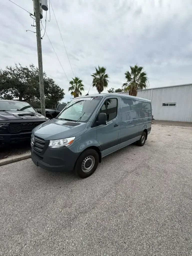 A gray Mercedes-Benz Sprinter van is parked on an asphalt lot under a cloudy sky. Palm trees and a white building are visible in the background, with a dark vehicle partially seen on the left.