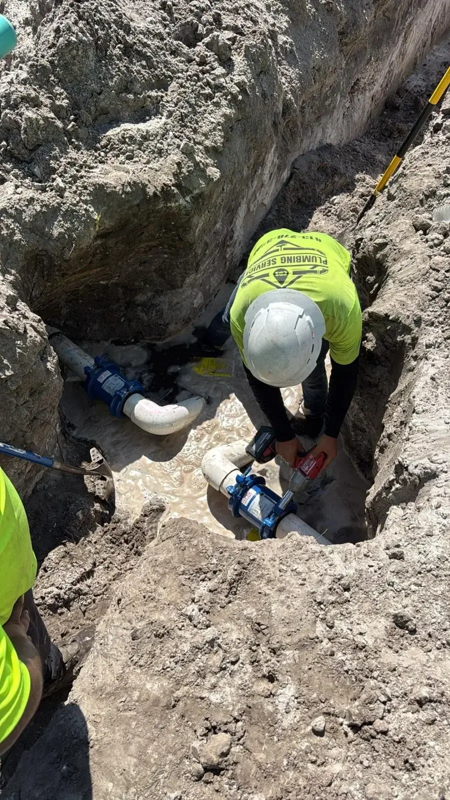 A plumber wearing a hard hat and a bright yellow 'PLUMBING SERVICE' shirt is working in a dirt trench, using a power tool on white PVC pipes with blue valves. The trench contains muddy water and loose dirt.