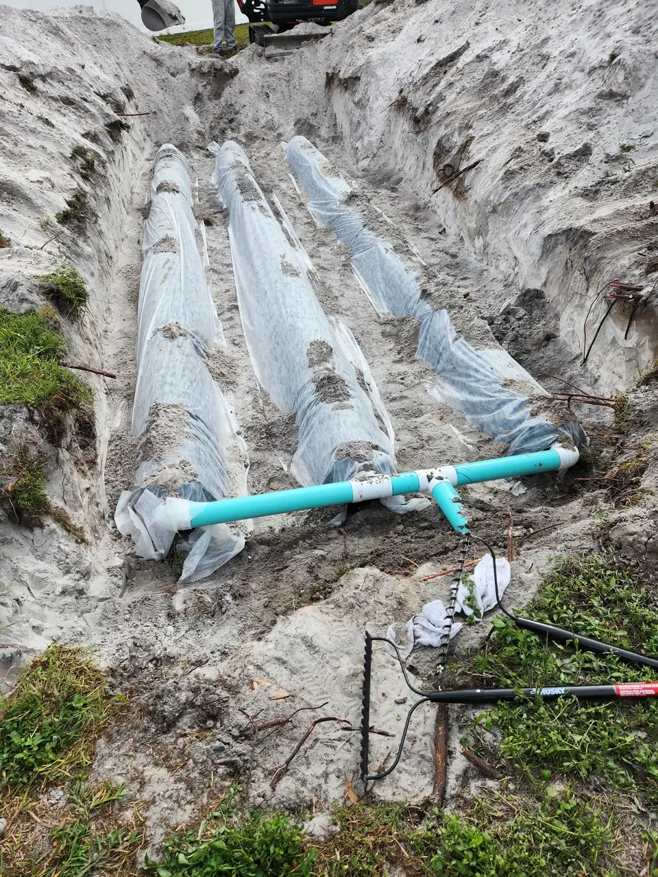 A trench dug in sandy soil contains three long, cylindrical objects wrapped in white fabric, partially covered with sand. A light blue PVC pipe assembly connects to one of the wrapped objects, with a person and an excavator visible at the top of the trench.