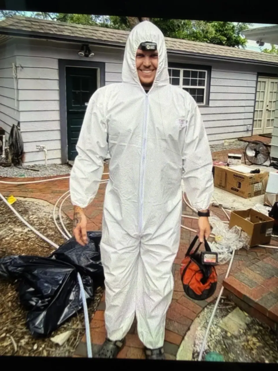 A man in a white hazmat suit with a headlamp smiles while standing on a brick patio, holding an orange and black bag. In the background is a house with light siding and various items like black trash bags, boxes, and a fan.